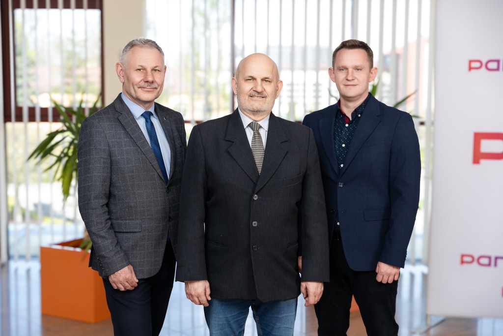 Trzech mężczyzn w garniturach pozuje do zdjęcia w nowoczesnym biurze.