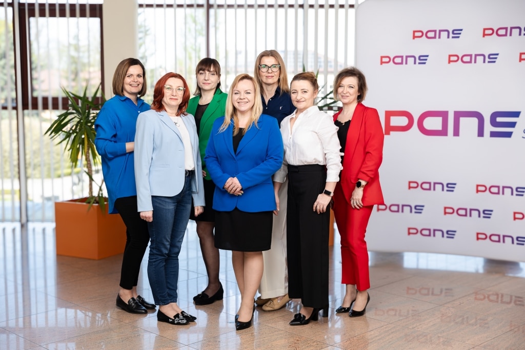 Zespół PANS w strojach biznesowych, siedem kobiet w różnym wieku, pozuje do zdjęcia w nowoczesnym biurze.