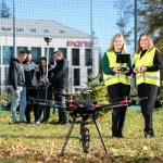 Geodeci z dronem i sprzętem pomiarowym na trawniku przed budynkiem.