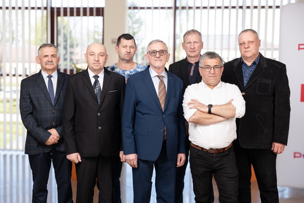 Grupa mężczyzn w garniturach i koszulach na tle żaluzji.