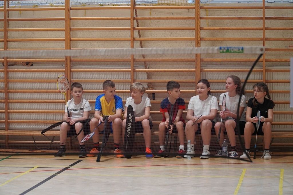 Dzieci siedzą na ławce w hali sportowej podczas turnieju badmintona WOŚP.