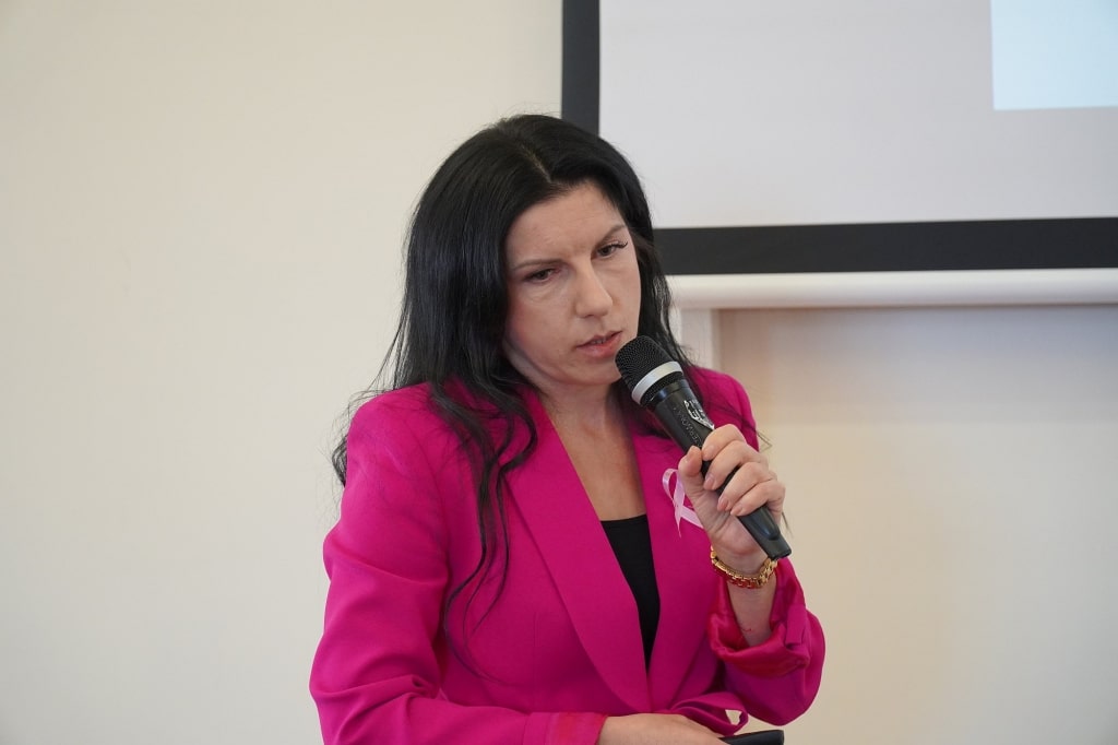Kobieta w różowej marynarce z różową wstążką mówi do mikrofonu na sympozjum
