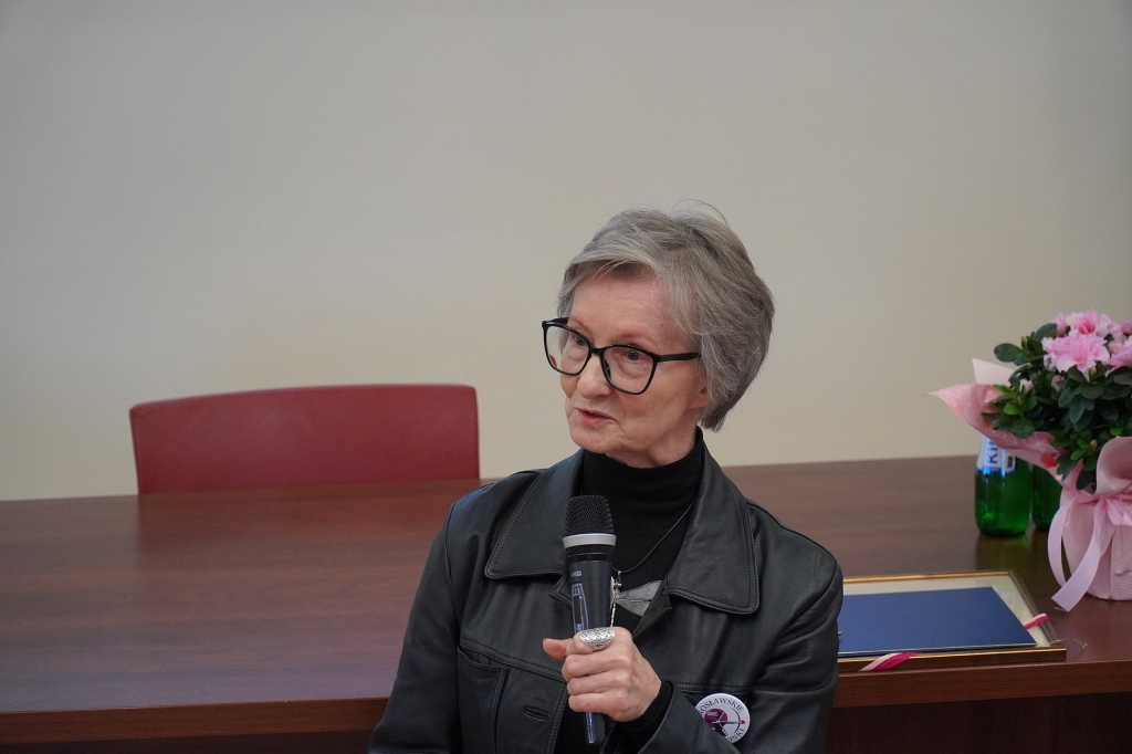 Kobieta w okularach mówi do mikrofonu podczas sympozjum Różowy Październik.