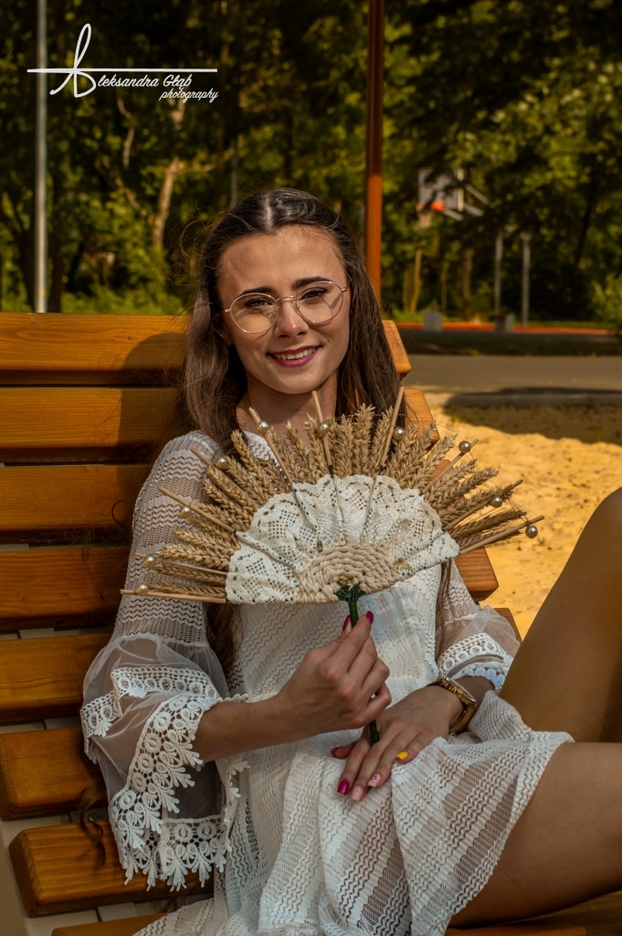Kobieta w białej sukience z wachlarzem z kłosów na ławce.