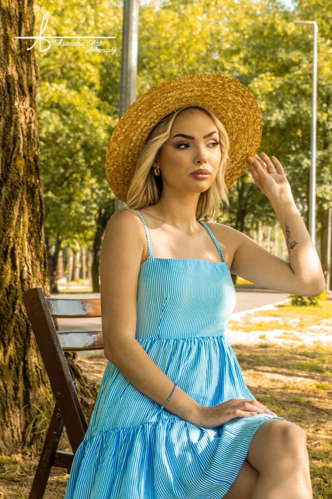 Kobieta w letniej sukience i kapeluszu siedzi na ławce w parku.