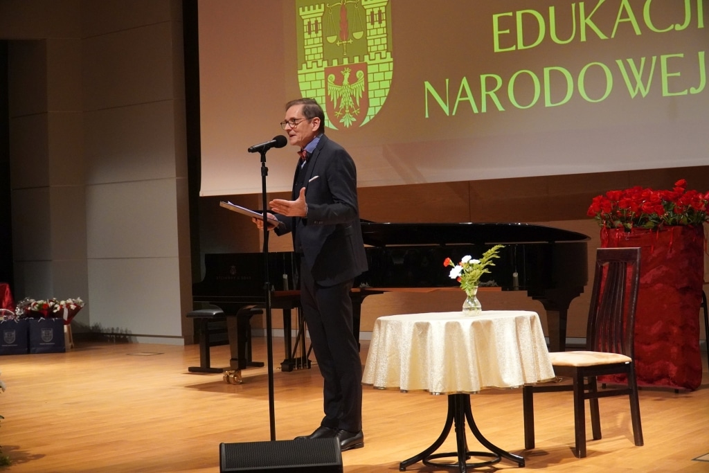 Mężczyzna przemawia na tle sceny z napisem „Edukacja Narodowa”