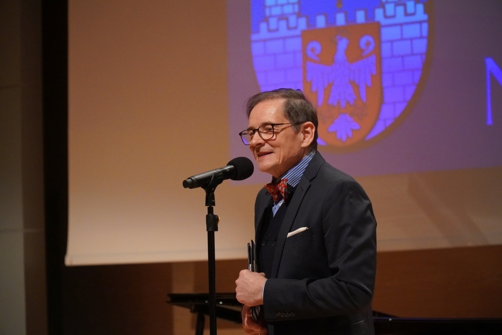 Mężczyzna w garniturze przemawia na tle herbu podczas Dnia Edukacji Narodowej.