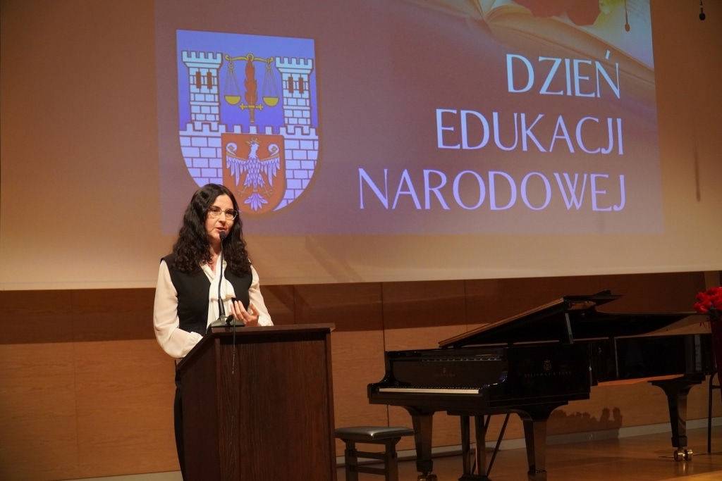 Kobieta przemawia podczas Dnia Edukacji Narodowej z herbem i napisem na ekranie.
