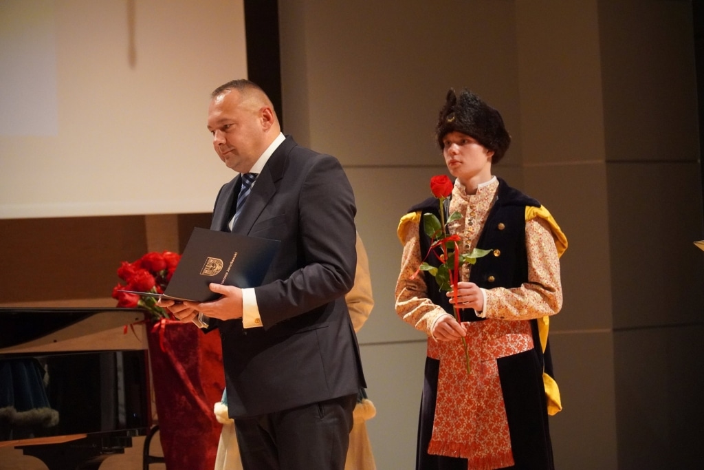 Uroczystość Dnia Edukacji Narodowej: mężczyzna z dyplomem i młody człowiek w stroju ludowym z różą.