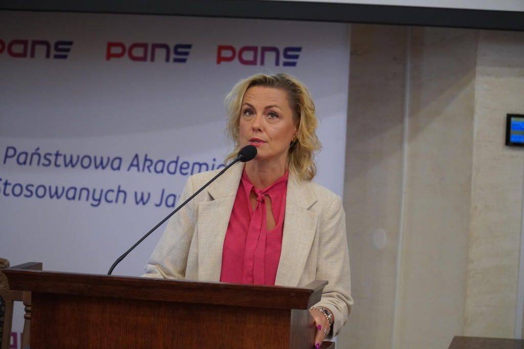Kobieta w różowej bluzce i beżowej marynarce przemawia przy mównicy