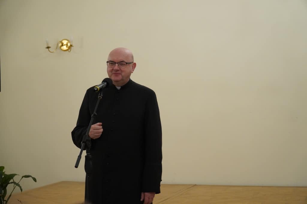 Ksiądz w sutannie przemawia na inauguracji roku akademickiego Akademii Trzeciego Wieku.