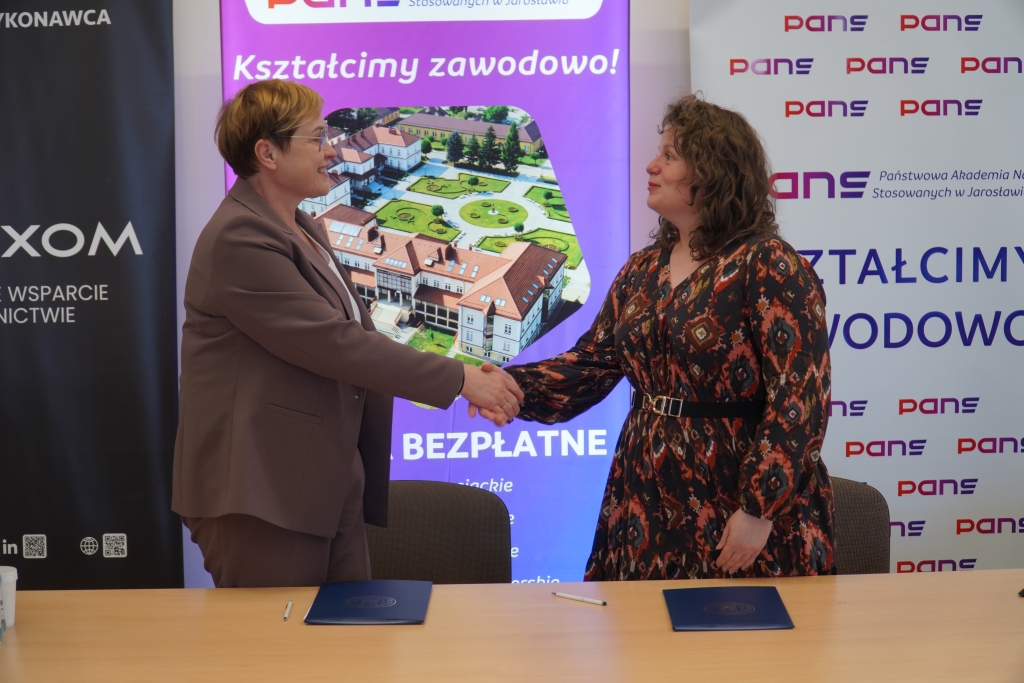 Dwie kobiety uśmiechają się, podając sobie ręce na tle baneru edukacyjnego.