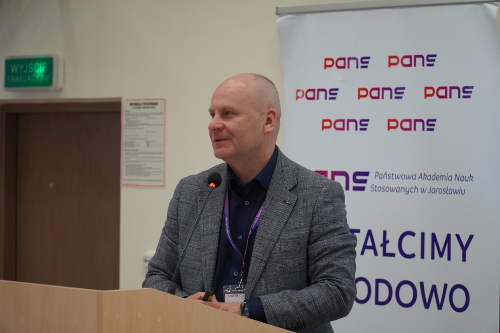 Konferencja naukowa WH: Mężczyzna w garniturze przemawia na tle logo PANS