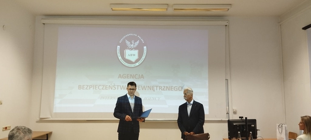 Spotkanie z delegaturą ABW: dwaj mężczyźni przed ekranem z logo Agencji Bezpieczeństwa Wewnętrznego.