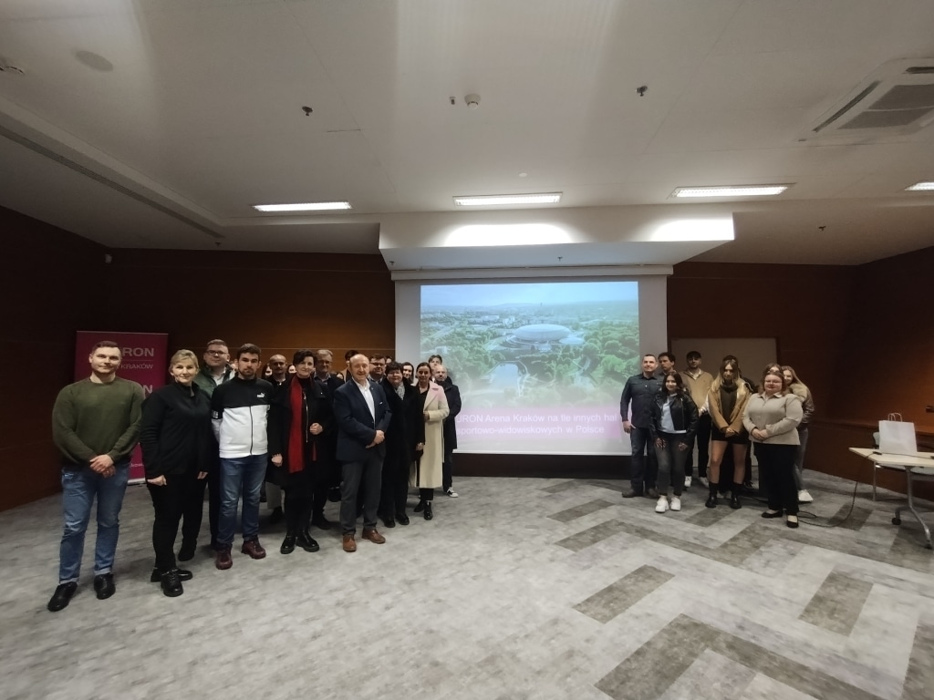 Grupa osób na tle sali konferencyjnej z projekcją TAURON Arena Kraków