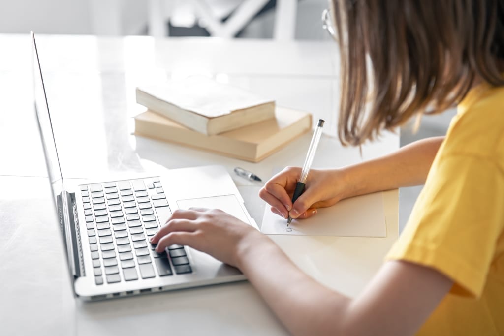Dziewczynka w żółtej bluzce uczy się online na laptopie, pisząc w zeszycie.