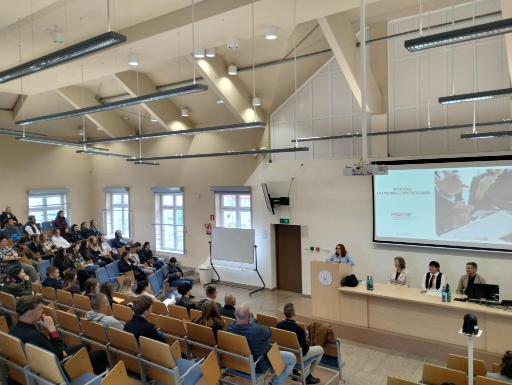 Spotkanie organizacyjne na uczelni PANS, prelegentka przy mównicy, studenci na widowni.