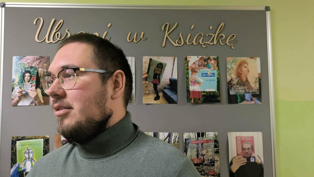Student psychologii na tle tablicy z książkami i napisem "Ubrani w Książkę" w SOSW Skawina.