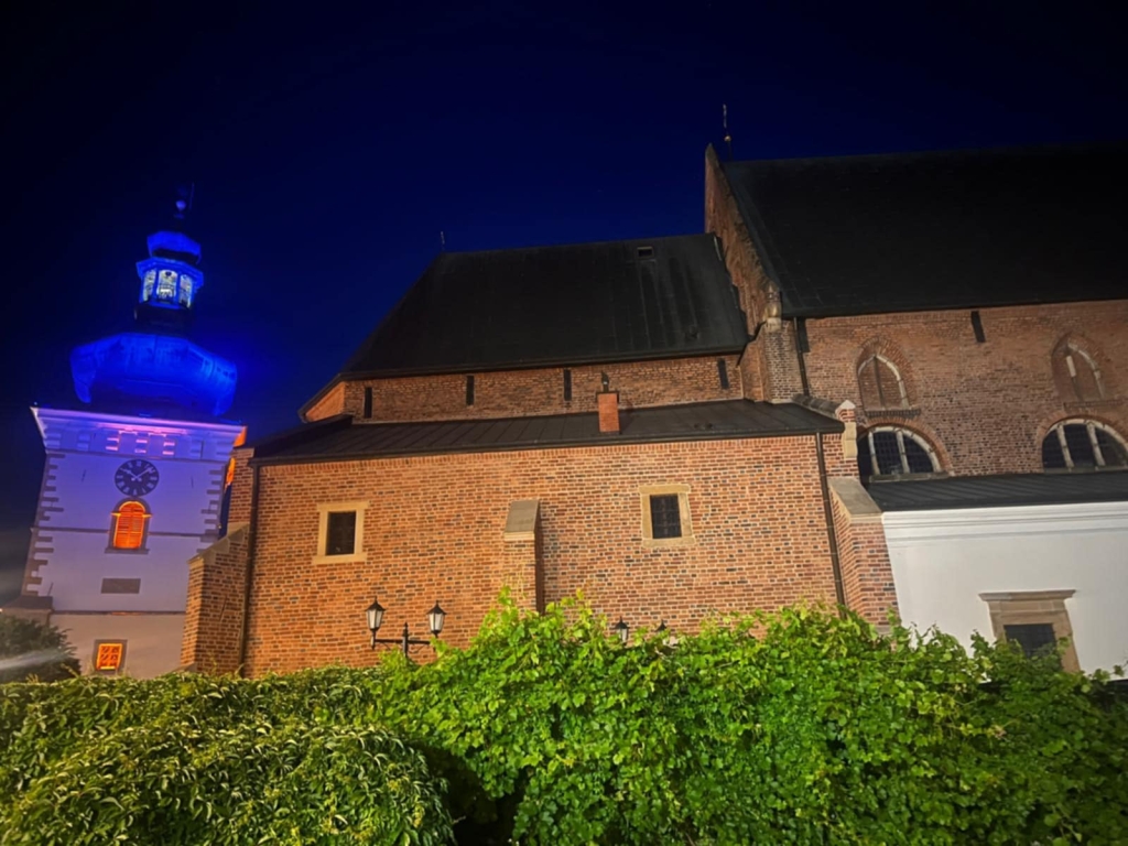Wieża kościelna oświetlona na niebiesko i pomarańczowo nocą, obok ceglanego kościoła.