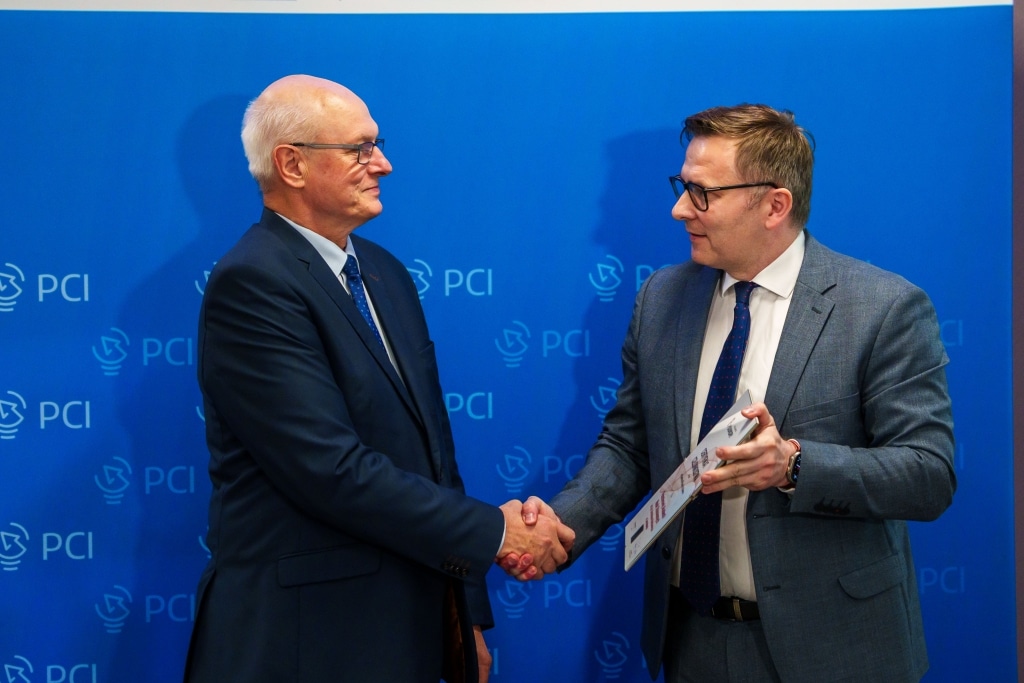 Dwóch mężczyzn w garniturach ściska sobie dłonie przed niebieskim tłem z logo PCI.