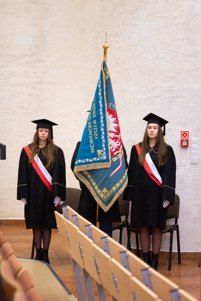 Inauguracja 2021/2022: dwie studentki w togach i biretach stoją przy sztandarze.