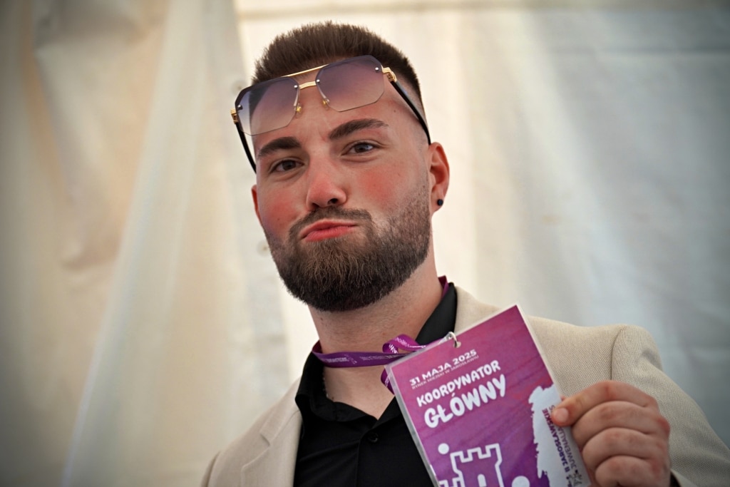 Młody mężczyzna w okularach trzyma identyfikator "Koordynator Główny" na Juwenalia 2025.