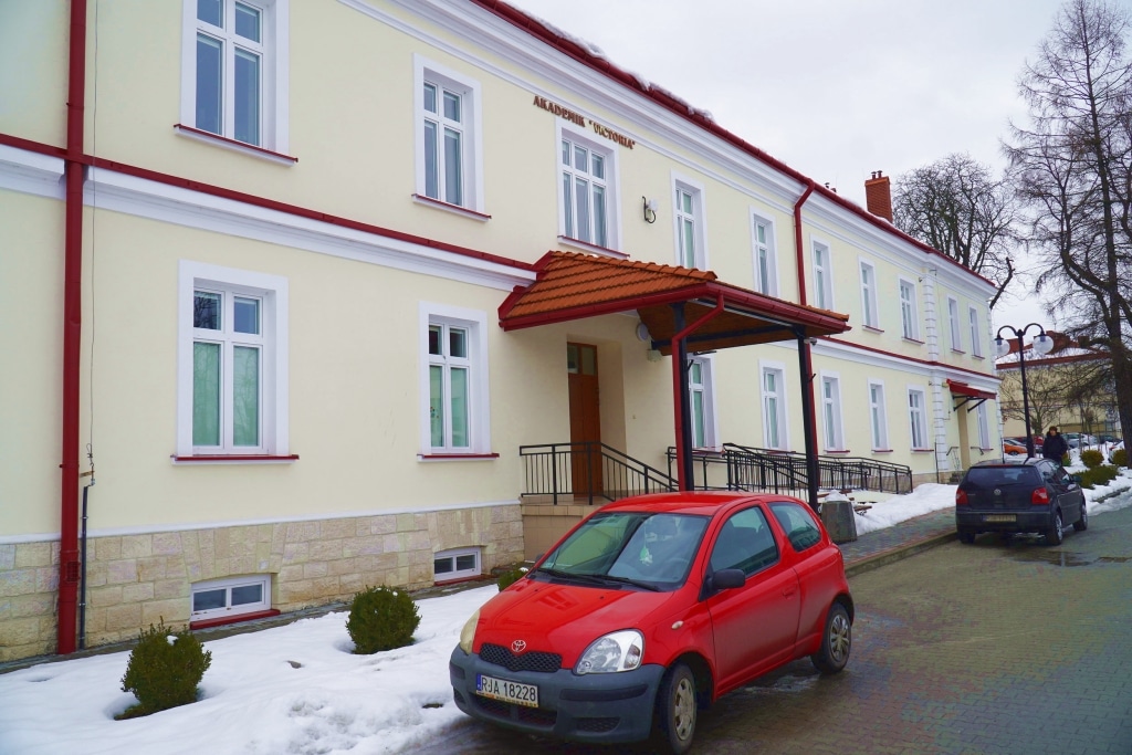 Czerwony samochód przed budynkiem Akademii PANS z napisem "AKADEMIA POLONIA" na fasadzie.