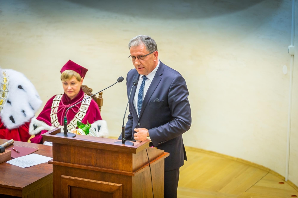 Mężczyzna w garniturze przemawia podczas Inauguracji 2018/2019, obok siedzi kobieta w todze.