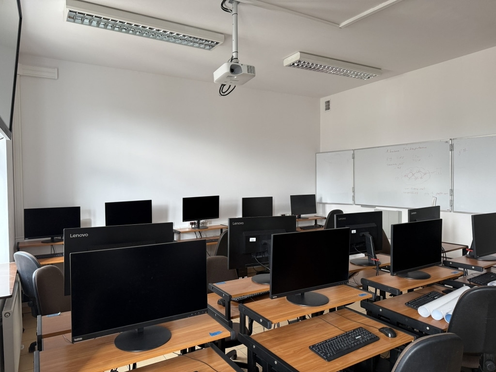 Nowoczesna sala komputerowa z rzędami stanowisk z monitorami i projektorem.